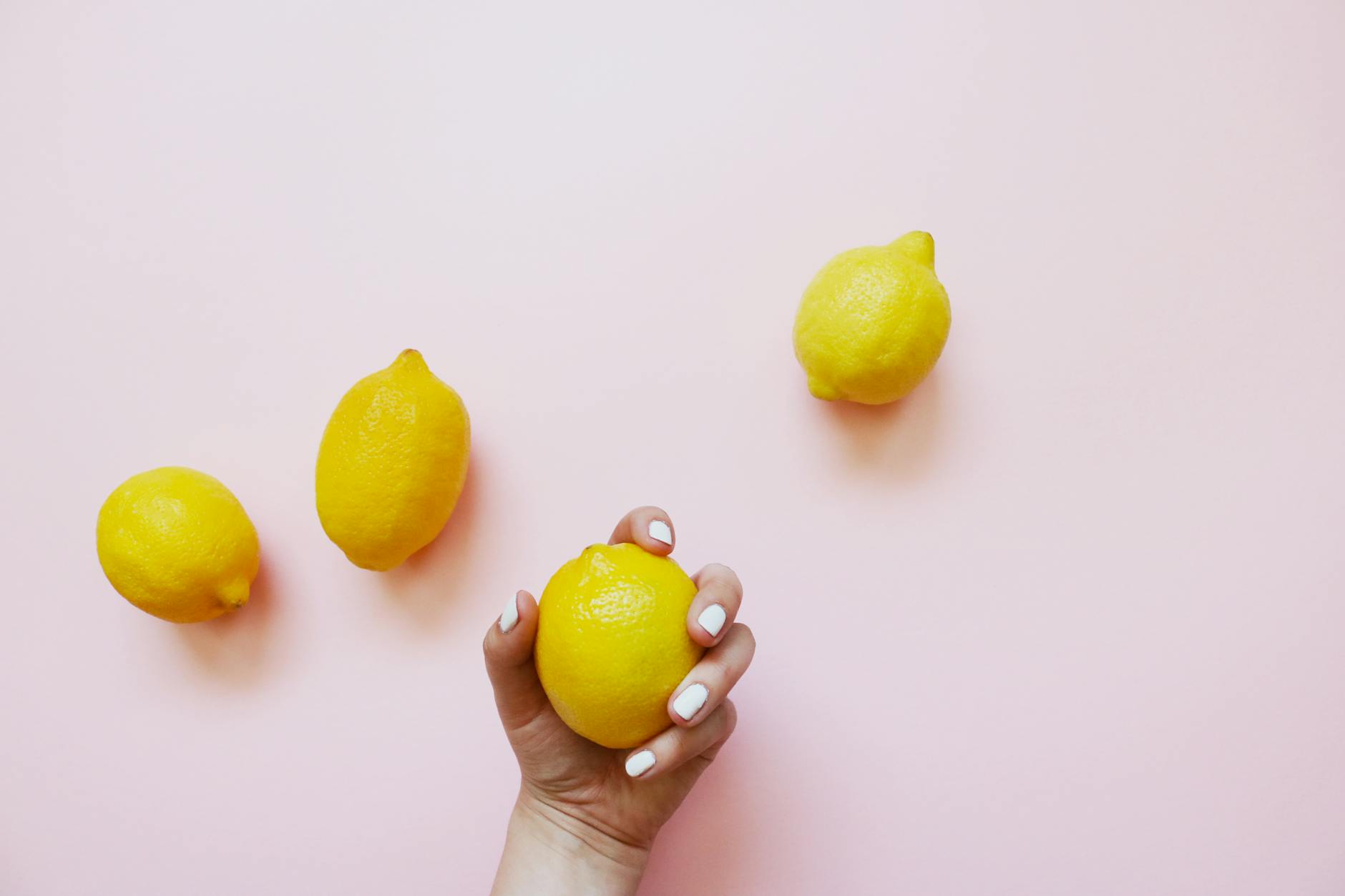 A hand with white nails holding a lemon on a soft pink background, surrounded by three additional lemons.