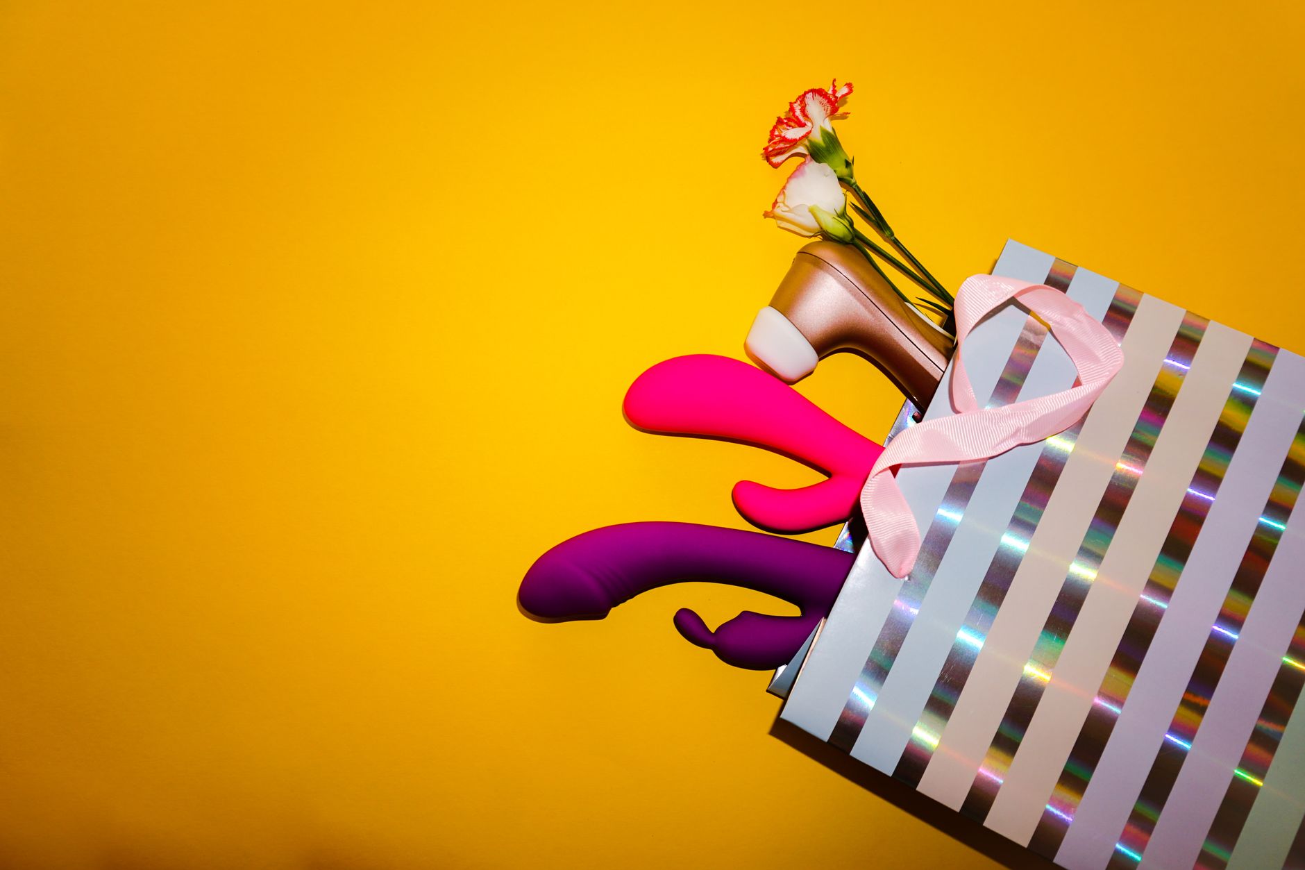 Colorful vibrators with flowers in a holographic gift bag on yellow background